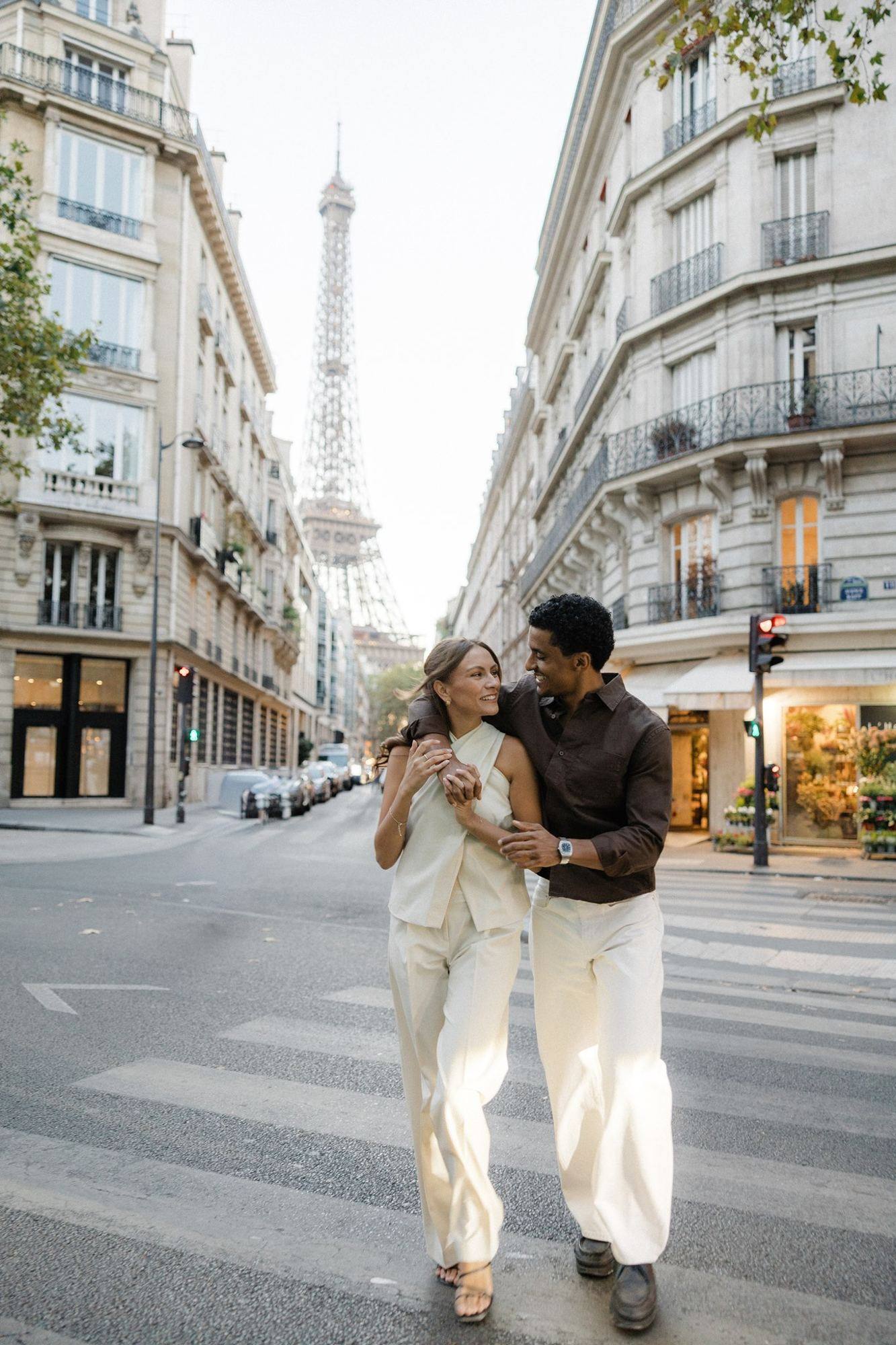 A couple crossing a Paris street, smiling at each other, the Eiffel Tower at the end of the street