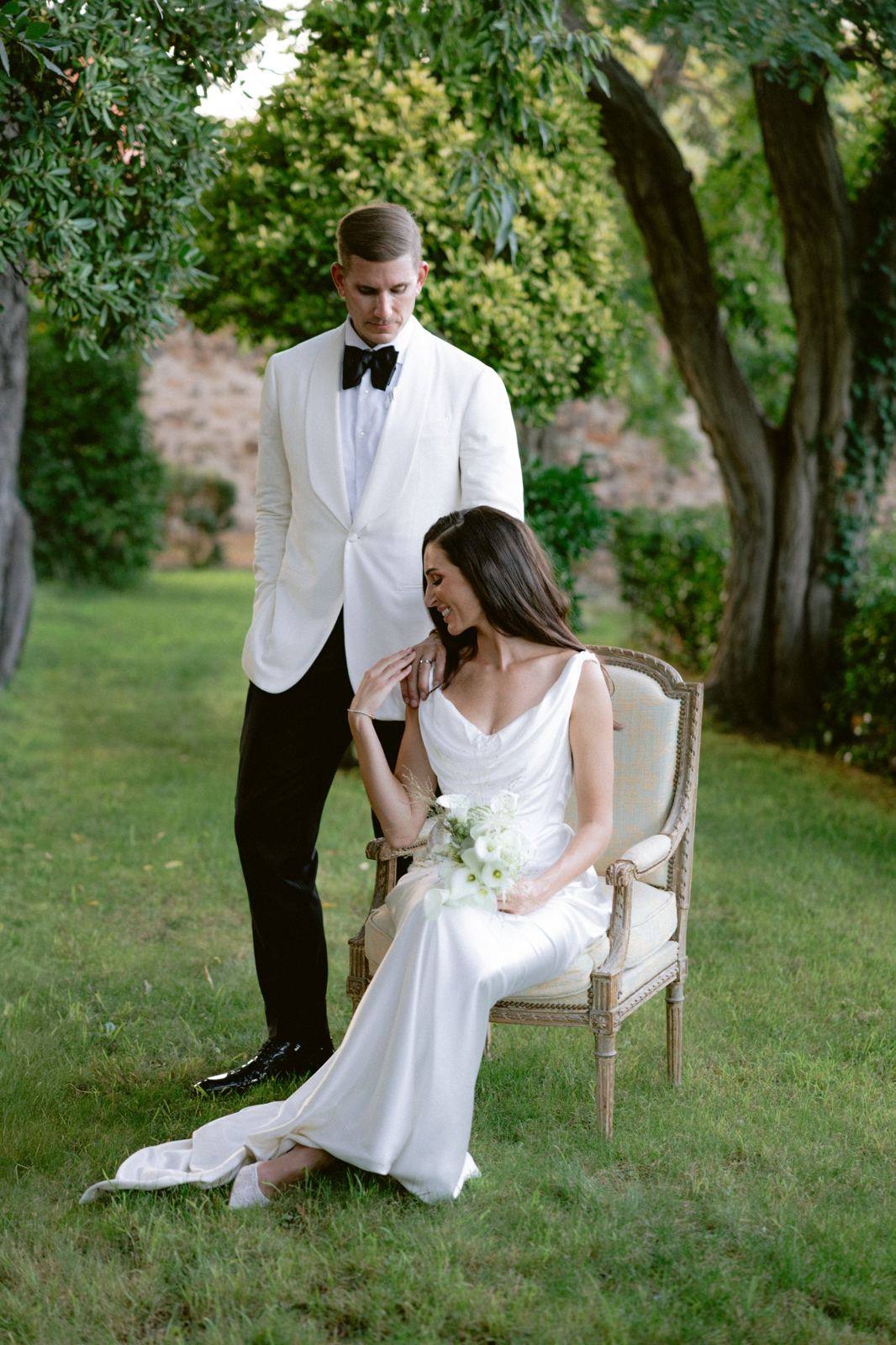 Bride seated in an armchair under the trees, the groom beside her