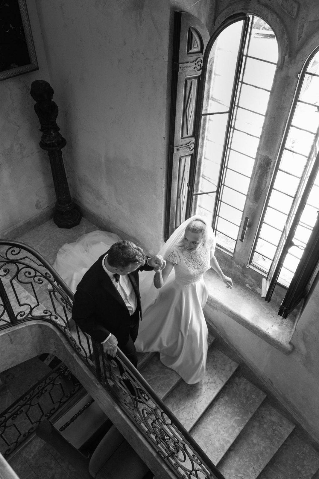 Bride walking down a stone staircase on the arm of a man in a tuxedo, by a tall arched window